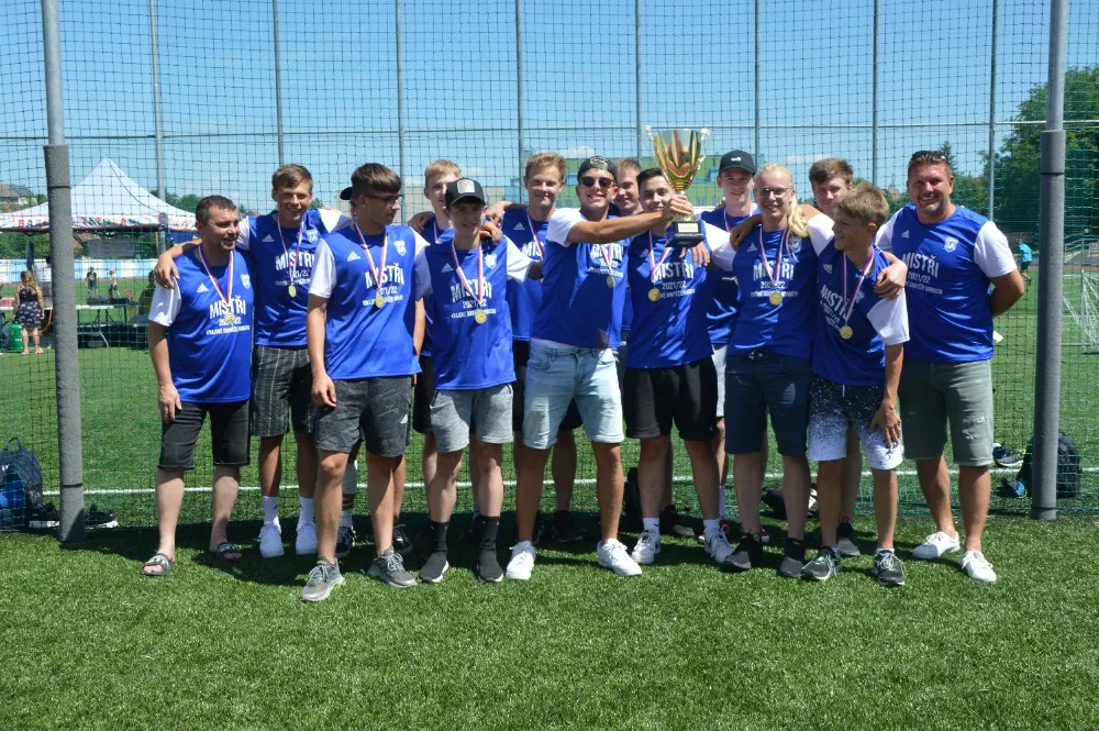 Hráči v modrých tričkách s trofejí na hřišti při oslavě 90 let kopané